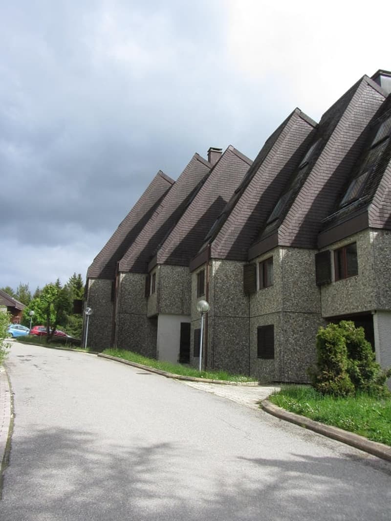 Multi-story apartment building with distinctive angular roofs, made of brick and stone, with parking spaces visible in the foreground
