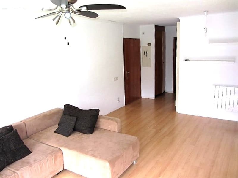 Spacious living room with hardwood floors, white walls, and a ceiling fan. The room features a beige sofa with black throw pillows. The entrance to the apartment is visible in the background.