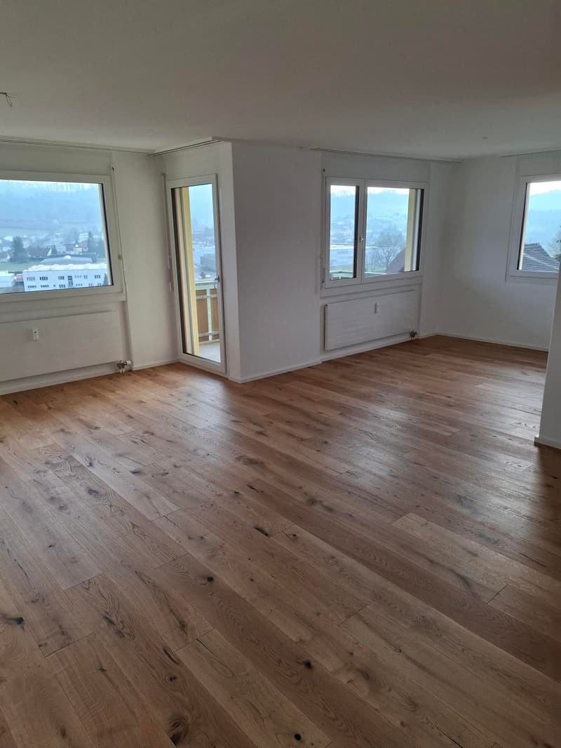 Spacious living room with large windows, hardwood flooring, and a sliding glass door leading to a balcony or terrace.