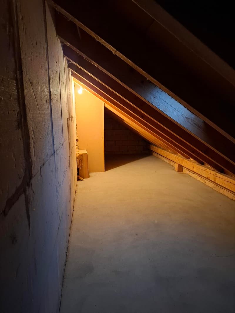 The image shows the interior of an attic space. The walls are made of wooden beams and the floor is covered in snow or insulation material. There is a single light source illuminating the space, creating a warm, cozy atmosphere.