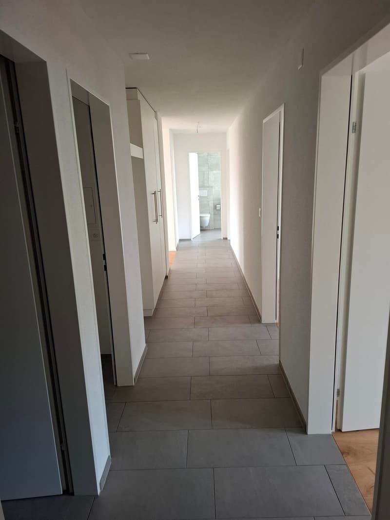 The image shows a long, narrow hallway with white walls and gray tile flooring. The hallway has several doors leading to other rooms, and there is a window at the end of the hallway providing natural light.
