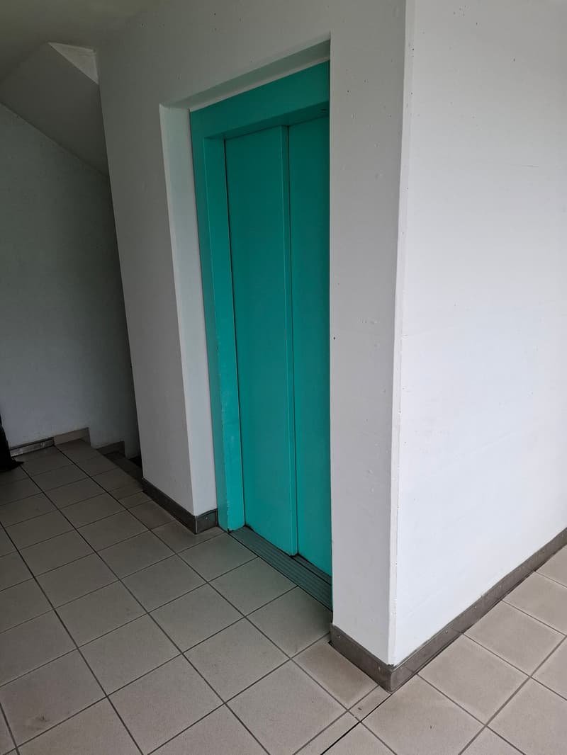 The image shows a hallway with a teal-colored door frame and white walls. The floor is covered with gray tiles.