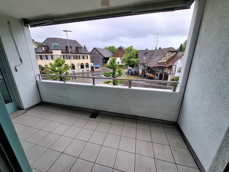 The image shows a balcony or terrace overlooking a small town or village. The view includes several buildings with a mix of architectural styles, including some traditional half-timbered houses. There are trees and greenery visible, and the scene appears