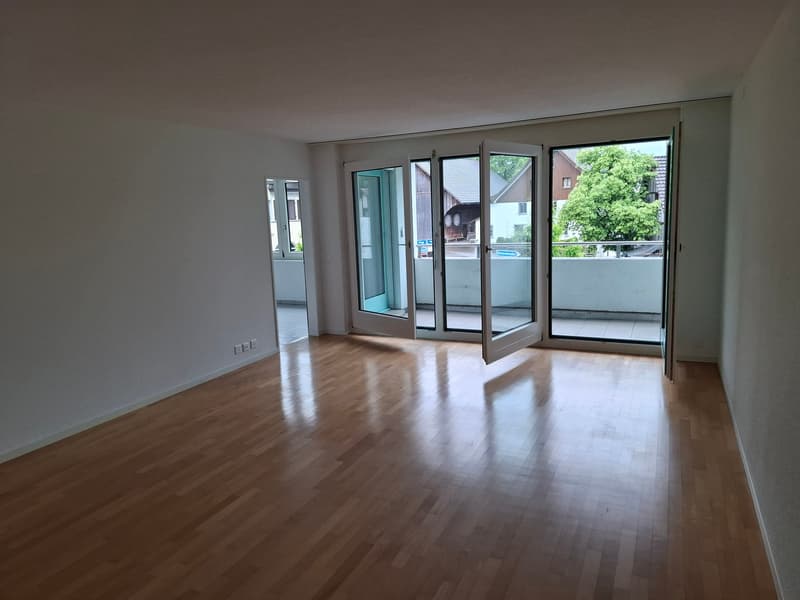 Spacious living room with hardwood floors, large windows, and access to a balcony or terrace overlooking the surrounding greenery.