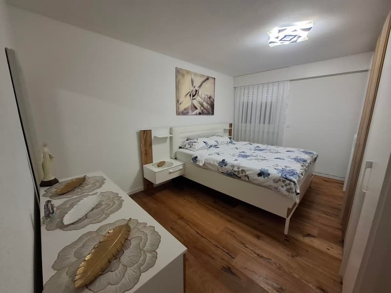 white walls, wooden floor, bed with white headboard, nightstand with drawers, decorative items, curtains