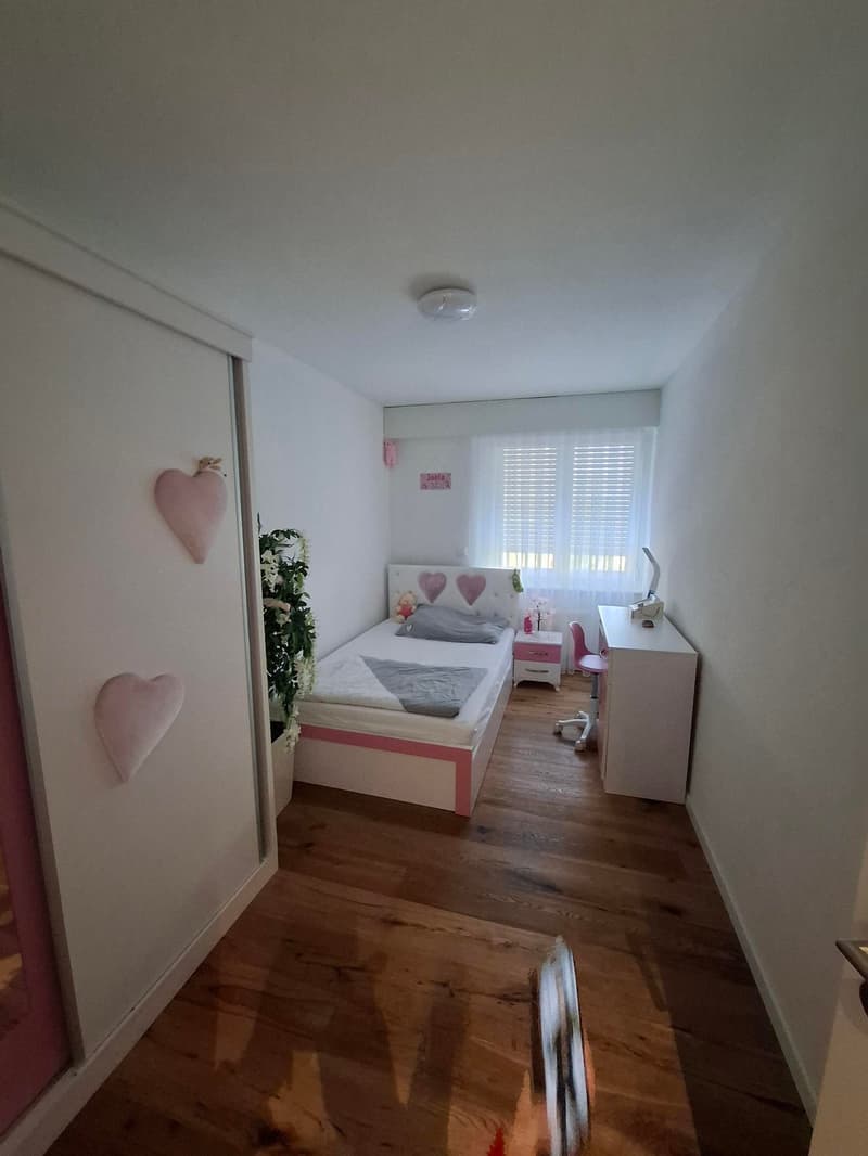 white walls, wooden floor, bed, nightstand, pink chairs, desk, potted plant, heart decor, window with white curtains