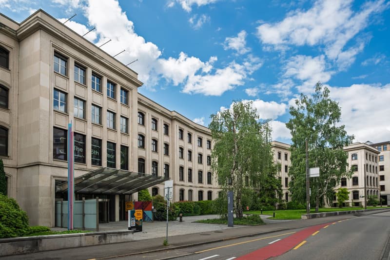 Large office building with multiple windows, beige facade, trees, street, grass, and parked bikes