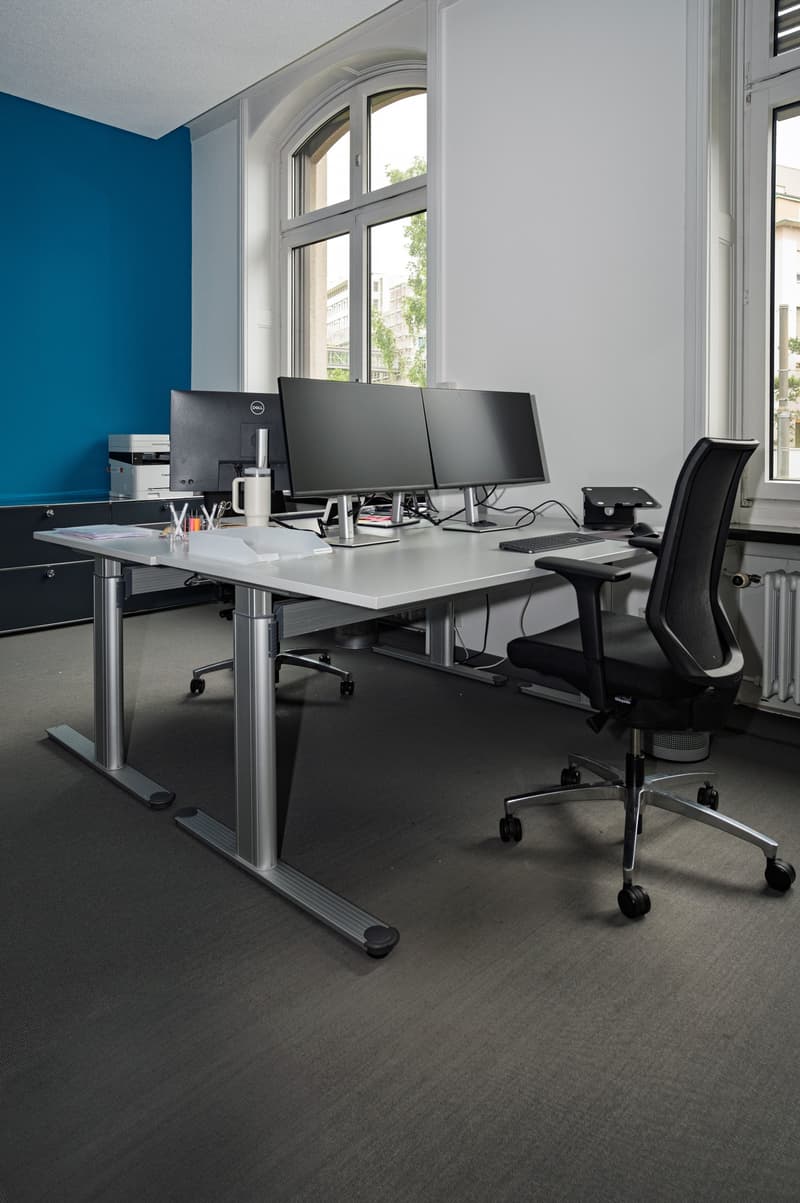 An empty office with an L-shaped white desk, two computer monitors, keyboard, mouse, black office chair, printer, two glass windows with trees outside, and a blue wall.