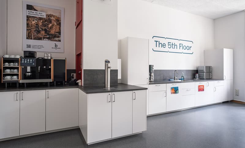a modern kitchen-lounge with various appliances, white cabinets, granite countertops, a large advertisement poster, a coffee maker, a sink with faucet, and a counter with a built-in coffee dispenser