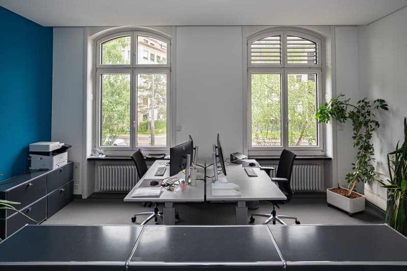 two desks with computers, office chairs, printer, plants, windows, radiators