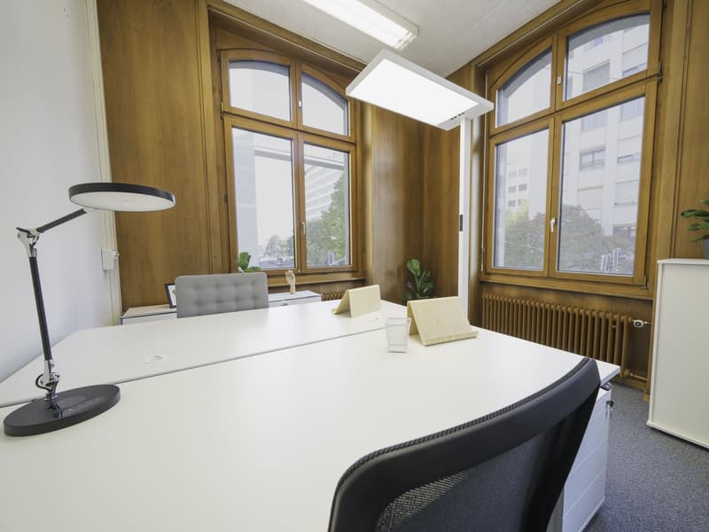 A room with a modern interior, featuring two white desks with office chairs, lamps, and office supplies. Wooden walls, large arched windows, and radiators are present. Outside the window, there is a view of the cityscape with buildings and trees.