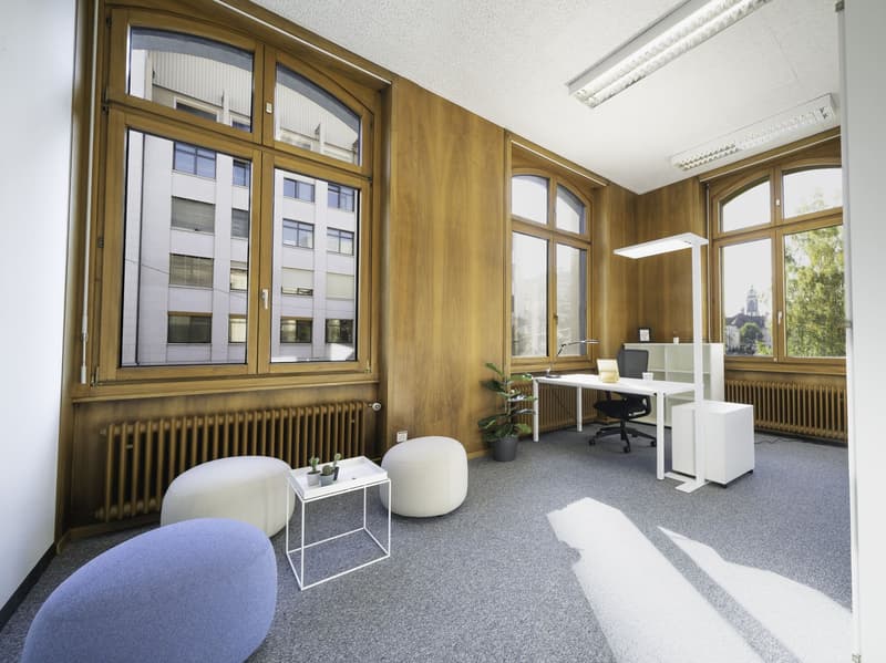 Contemporary office space with wooden panels, large windows, desk, monitor, lamps, radiators, and seating area with bean bags