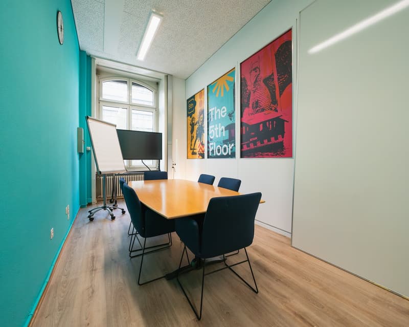 Office room with wooden desk, blue chairs, whiteboard, monitor, wall paintings, clock, windows, radiator, power outlets, and wooden flooring.