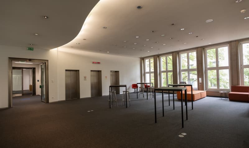 An empty open area, several black tables, a couple of chairs, a couch, three elevators, white walls, large windows, ceiling lamps, carpeted floor.