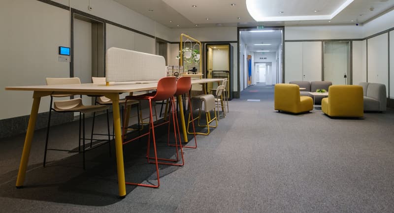 A room with a long table with red and yellow chairs, a white desk, yellow armchairs, and a carpeted floor.