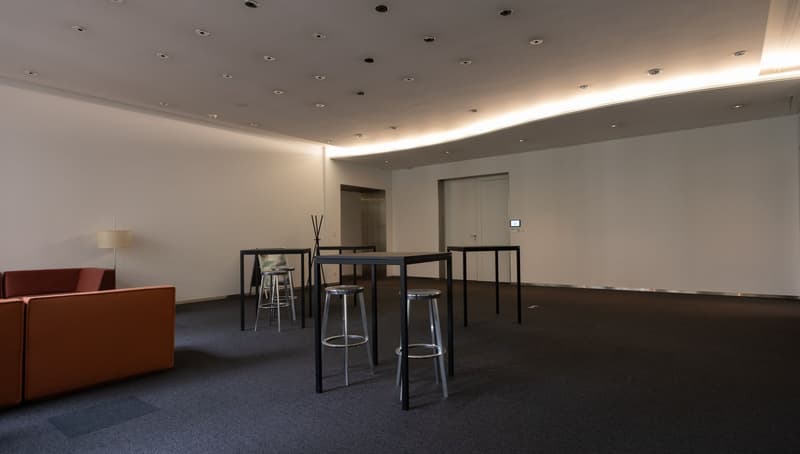 A room with a long table, chairs, lamp, brown couch, and white walls.