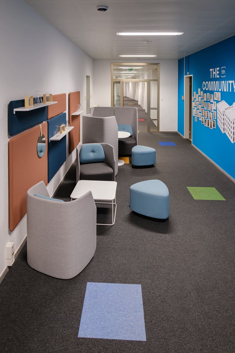a modern lounge with blue and gray chairs and ottomans, a blue wall with artwork, a white table, and gray carpet