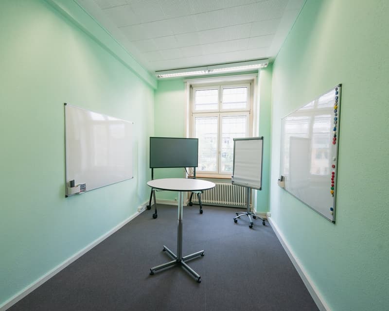 Empty room, green walls, whiteboards, central round table, movable whiteboard, flatscreen monitor on the wall, window with a view to the outside