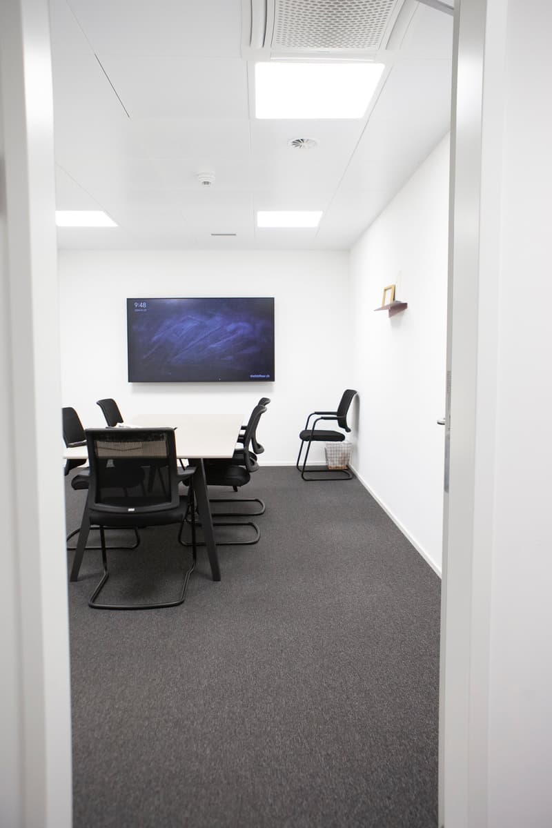 A meeting room with white walls, a ceiling with lights, a monitor mounted on the wall, a desk, and several black chairs.