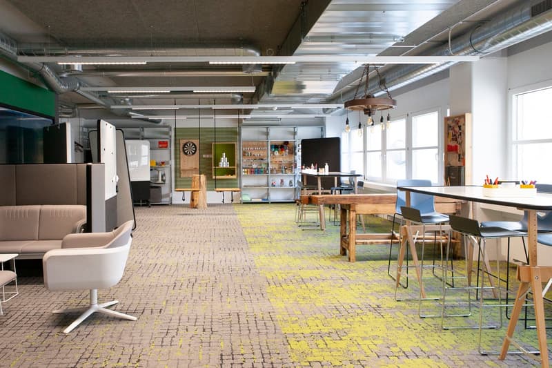 Modern office space with tables, seating areas, large windows, and utility shelves with various items.
