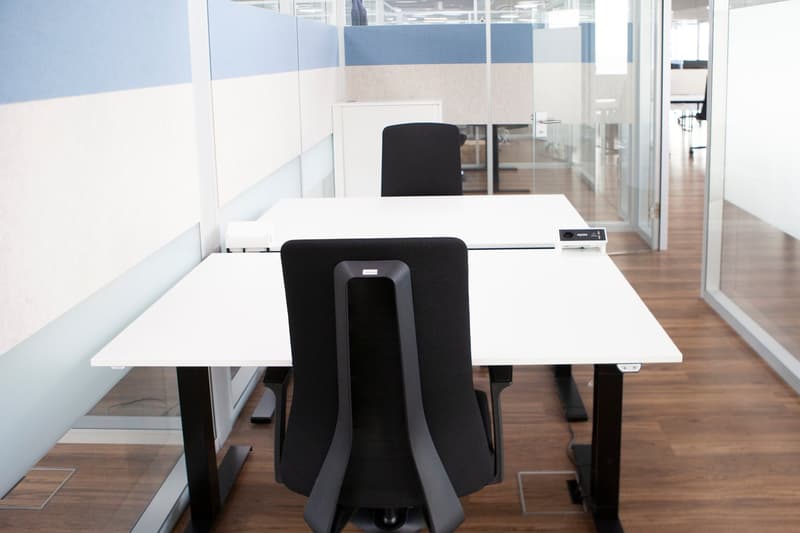 Two desks, glass walls, two black chairs, one of them adjustable, wooden floor.