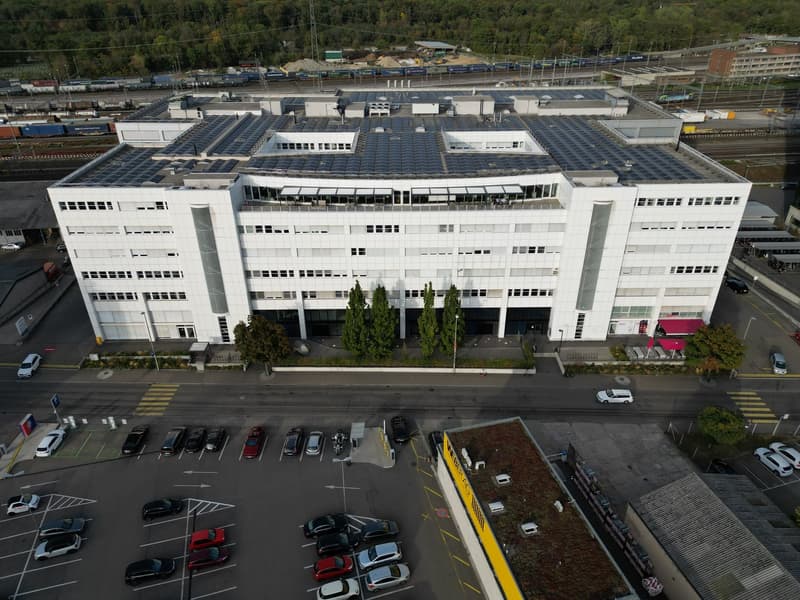 Large white building, many windows, solar panels on the roof, parking lot with several cars parked, surrounding trees, and a red awning on the right side
