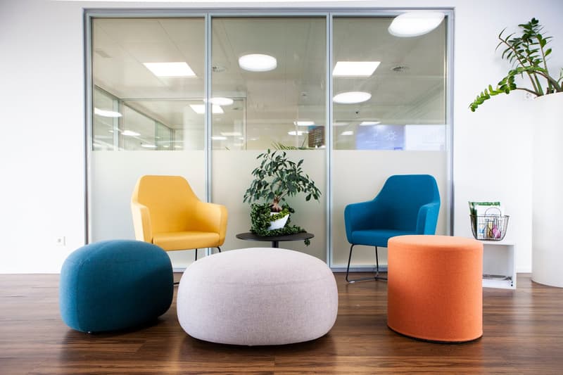 large room with white walls, glass partitions, round and square ottomans, 3 chairs, a center table with a potted plant