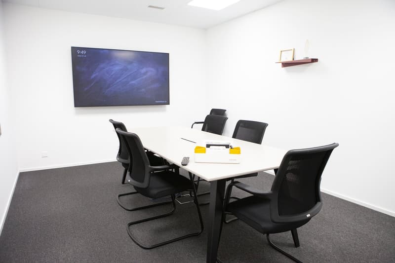 a blank meeting room with a large monitor on wall, a white desk and 6 black chairs, a frame on the shelf