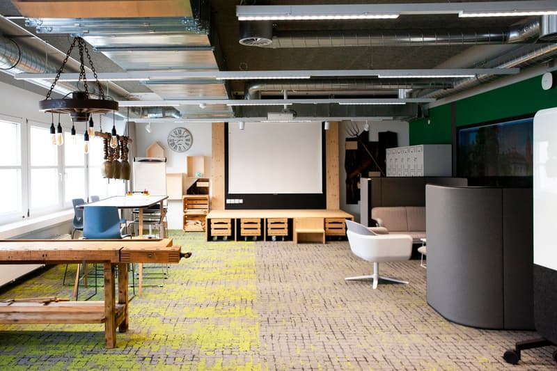 modern open office layout with wooden desks, chairs, meeting areas, a projector screen, decorative items, storage cabinets, green and white walls, suspended ceiling with lighting and ventilation