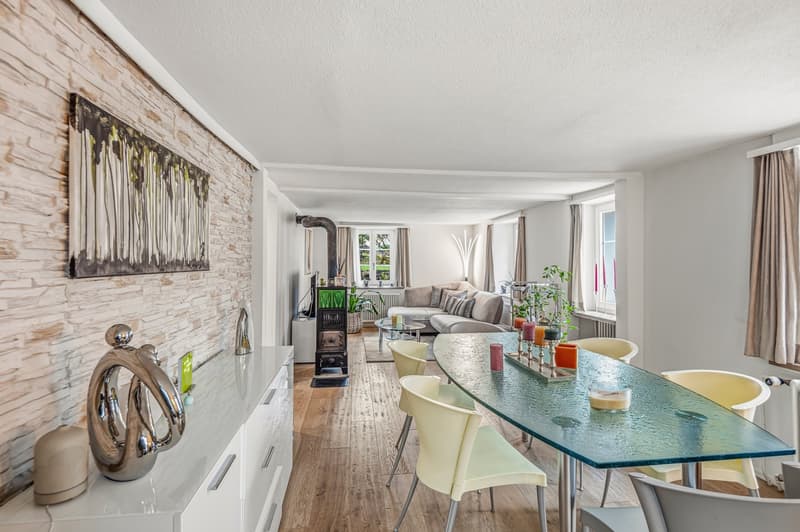 A spacious living room with brick wall, white painted walls, a sofa, coffee table, glass table, and chairs. It has a fireplace and white furniture. There is also a brick chimney, a white cabinet, and a glass top table.
