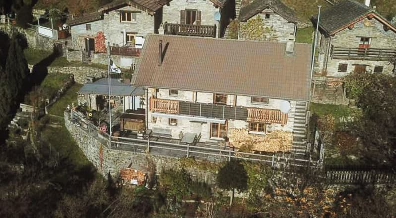 2 story stone house with brown roof, surrounded by other houses, balcony, garden, stone wall, trees