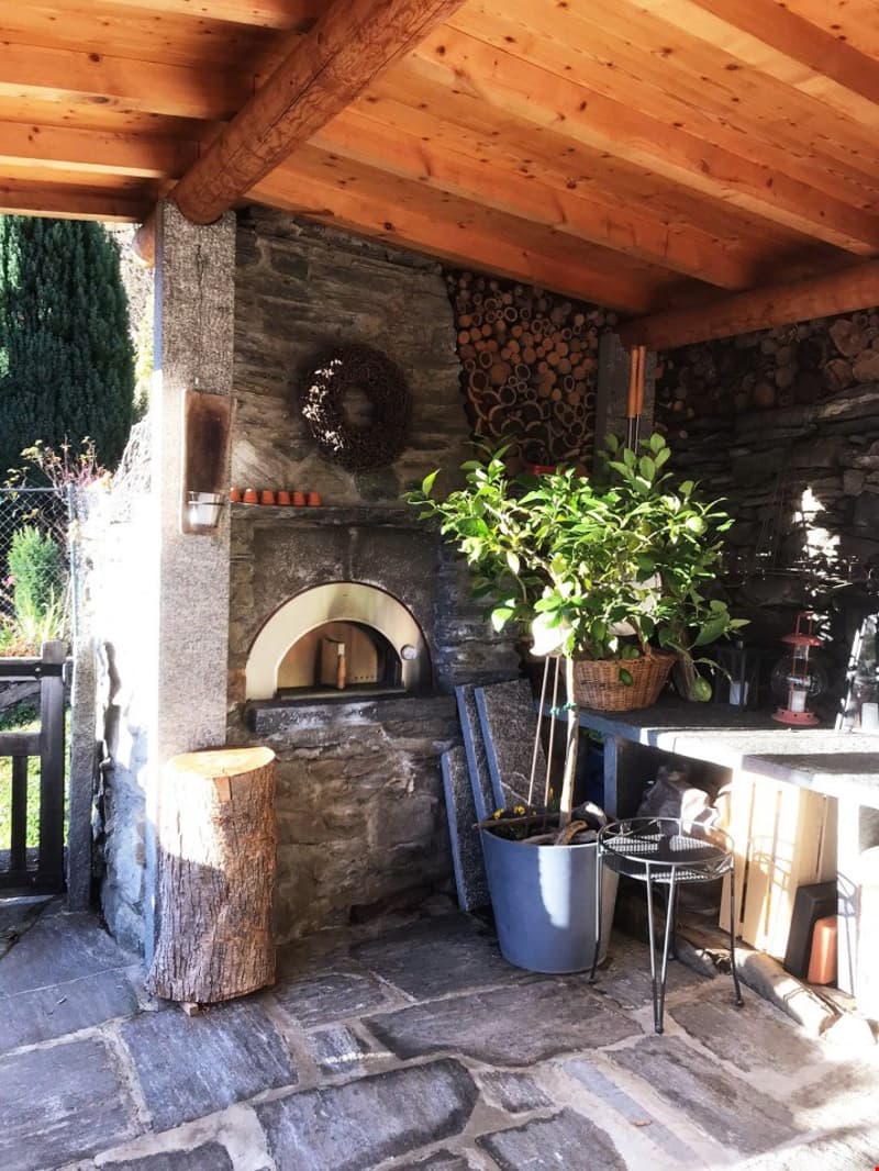 Outdoor kitchen with brick oven, wooden beams, stone flooring, potted plants, table, lamps, and stack of wood logs