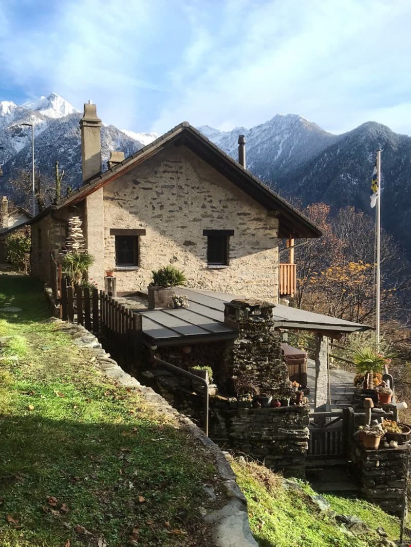 a stone house with a wooden front porch, elevated ground floor, chimney, windows, flag, and lush greenery, nestled against a mountainous backdrop