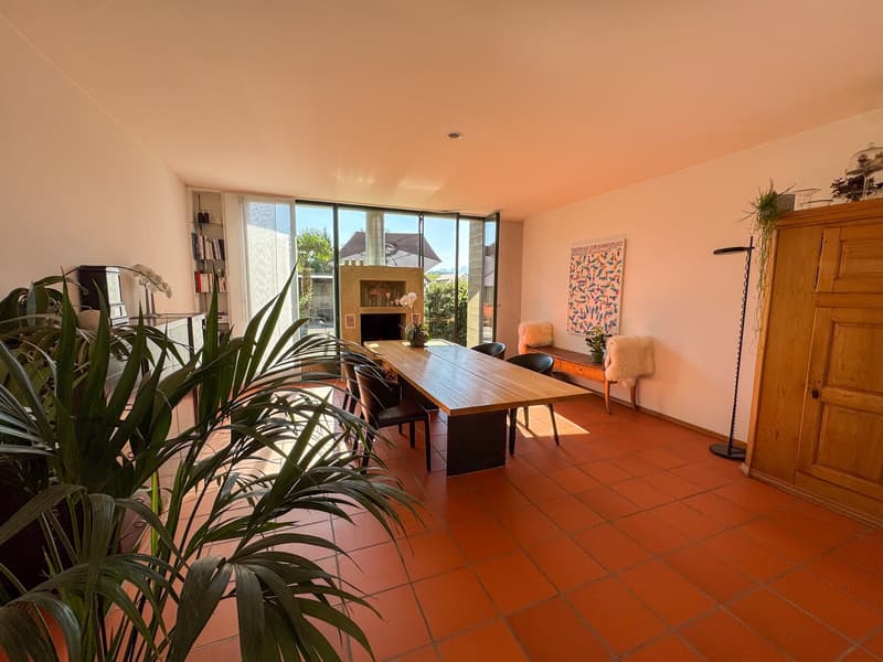 Long dining table, black chairs, orange tiled floor, large windows, fireplace, plants, artwork on wall, wooden cabinet, bench, potted plants