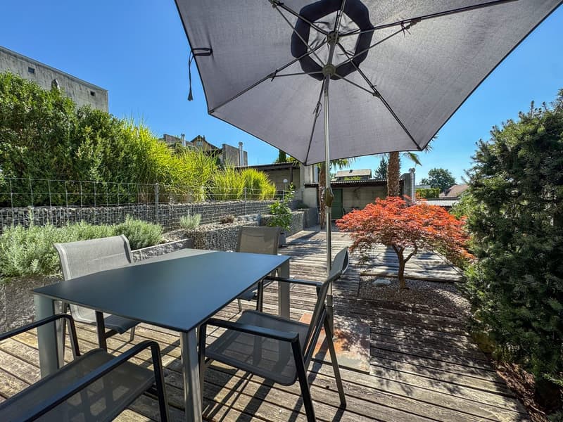 Wooden deck with a table and chairs, umbrella, surrounded by a fence with plants, building and trees in the background