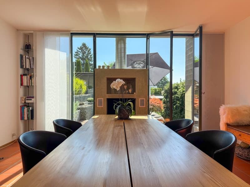 Wooden table, six black chairs, floor-to-ceiling windows, fireplace, potted flower on table