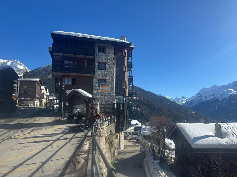 Mountainside building with multiple floors, stone and wood exterior, snow on the roof, surrounded by snowy mountains