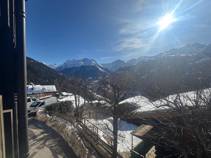 Mountains covered in snow, clear blue sky, houses with snow on roofs, trees without leaves, road with parked cars