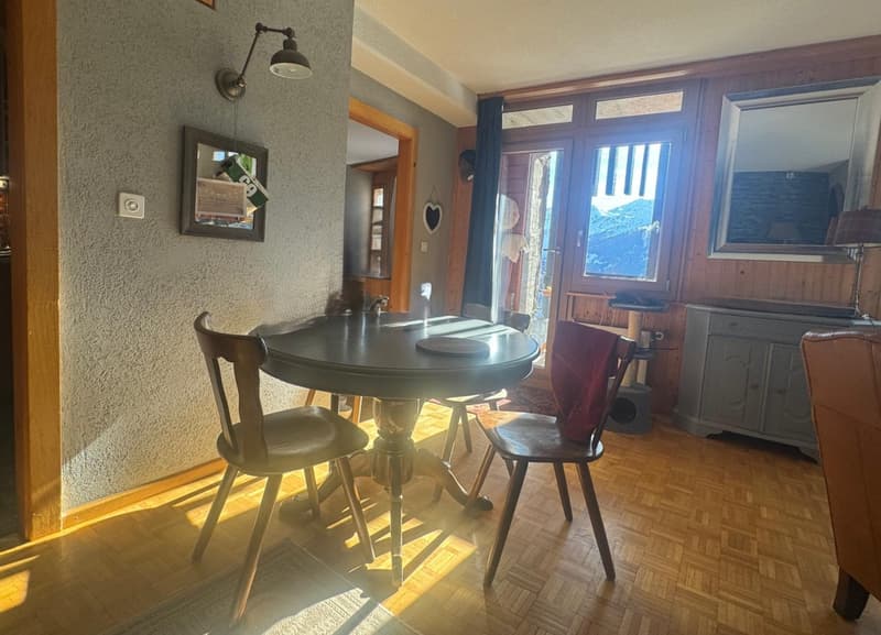 Dining area with a round table, wooden chairs, wooden floor, wall-mounted lamp, mirror, and a cabinet with a lamp on top.