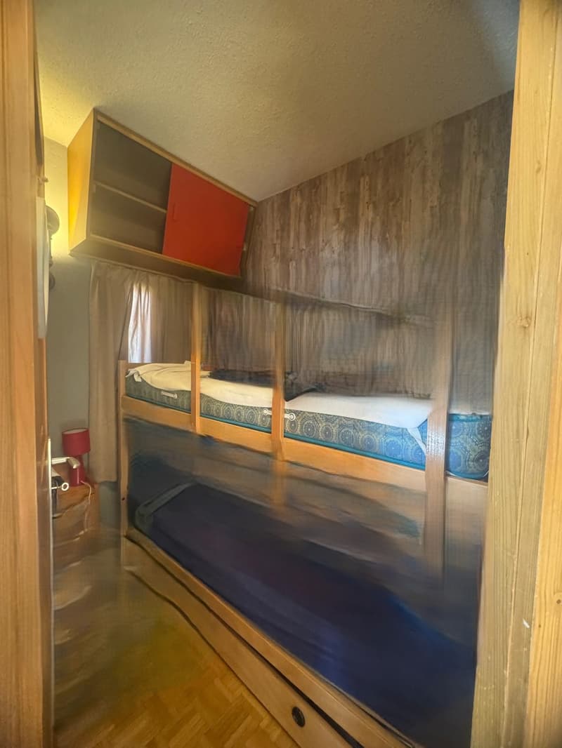 A room with wooden walls and floors, a bunk bed with three levels, a wooden cabinet with a red door, a lamp, and a curtain