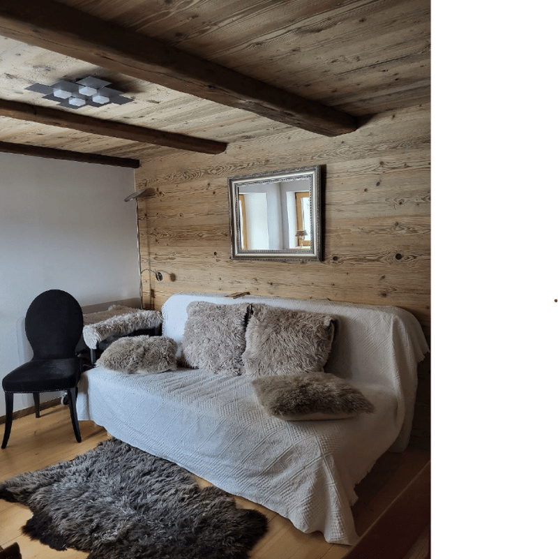 Wooden ceiling, a sofa with cushions and a rug in front of it, a black chair next to the sofa, a mirror mounted on the wall, and a small lamp inside the mirror