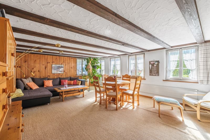 Wooden beams, wooden flooring, sectional sofa, dining table, dining chairs, coffee table, wooden cabinet, radiator, windows with white curtains, picture frame, potted plant