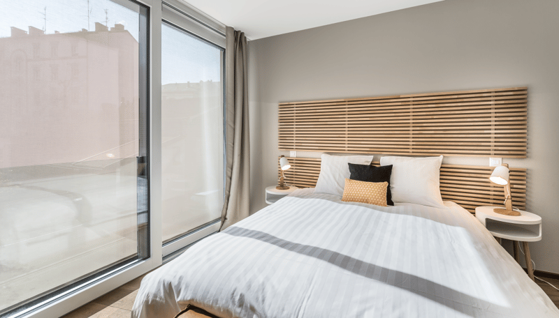 Spacious bedroom with large windows, wooden slatted headboard, white bedding, side tables, and lamps