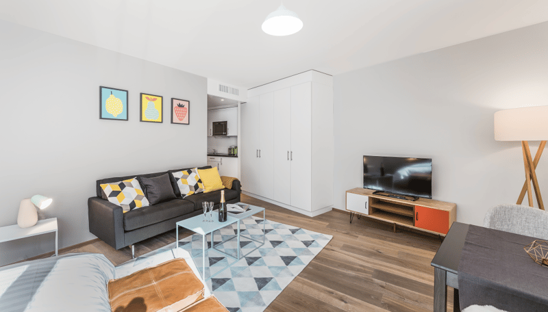 Spacious living room with hardwood floors, gray sofa with patterned cushions, coffee table, TV stand, and colorful artwork on the walls. The room also features a built-in wardrobe and a kitchenette area.