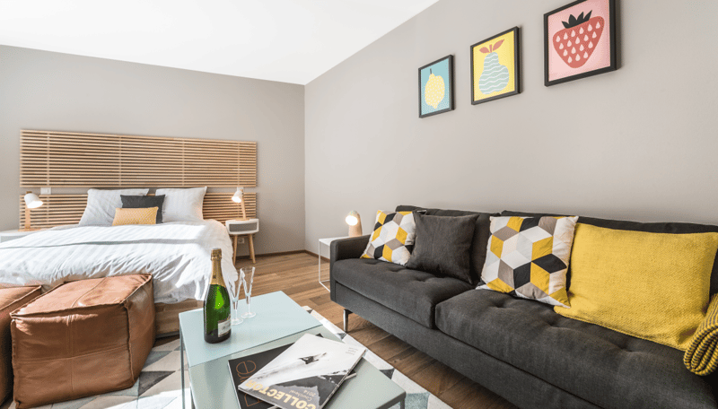 Open-plan studio apartment with a bed, sofa, coffee table, and decorative wall art. The room has a wooden headboard and shelving unit, and the walls are painted in a neutral color. There is a balcony or terrace visible through the window.