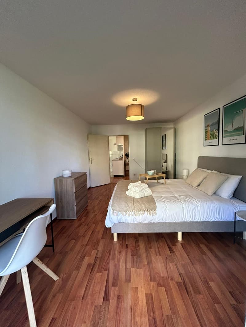 spacious bedroom with wooden floor, white walls, large bed with gray headboard, nightstand, wooden desk with chair, open door leading to kitchen, two paintings on wall, mirror