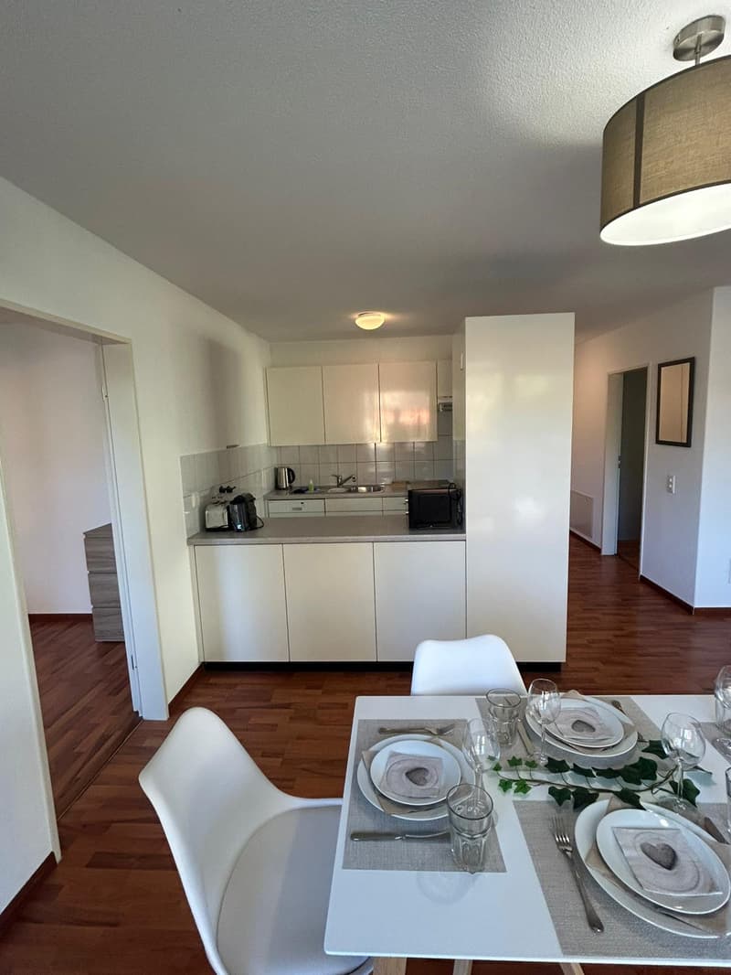 Dining area with dining table, white chairs, modern kitchen, white cabinets, sink, dishwasher, microwave, refrigerator, toast, plate, glass, wooden floor