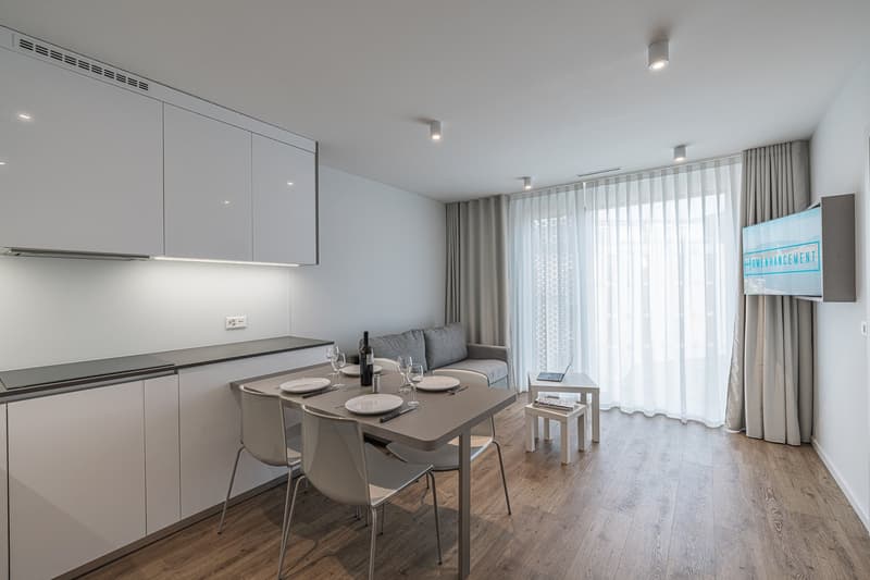 Modern, minimalistic open-plan living space with dining table, modern kitchen, grey sofa, TV, and large windows with curtains