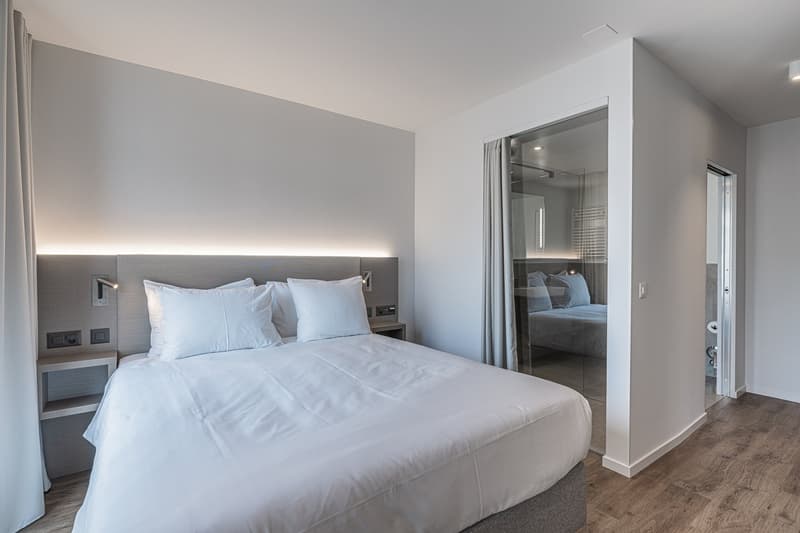 a room with a bed, nightstands, bedside lamps, gray headboard, glass door, white walls, windows, wooden floor