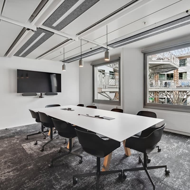 A conference room with a white long table, black swivel chairs, large windows, large flat screen TV mounted on the wall, gray carpeted floor.